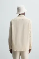 Sobrecamisa de hombre de corte relajado, color beige claro con finas rayas verticales en tono más oscuro. Presenta cuello bowling, manga larga con puño abotonado, bolsillos de parche en el pecho y en la cadera, y aberturas laterales en el bajo.