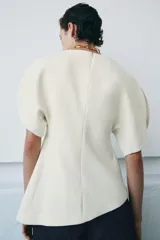 Top color crema de cuello redondo y manga corta abullonada. Presenta un bajo asimétrico y cierre en la espalda con cremallera oculta en la costura.