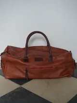 Bolso de viaje tipo weekender de cuero color tostado con asas y detalles en marrón oscuro, herrajes metálicos y bolsillo frontal con cierre.