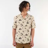 Camisa de manga corta color beige con estampado tropical de palmeras y veleros, cuello abierto y bolsillo en el pecho.