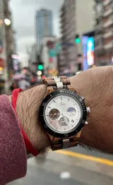 Reloj cronógrafo con caja de madera de zebrano y bisel técnico de acero negro. La esfera es plateada con subdiales de madera, indicador de fase lunar y fechador. La malla combina eslabones de acero inoxidable con madera de zebrano.
