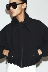 Chaqueta bomber negra con cuello redondo y pañuelo desmontable anudado al frente. Presenta cierre frontal con cremallera, bolsillos laterales con solapa y botones a presión, y terminaciones elásticas abullonadas en puños y ruedo.