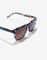 Lentes de sol Hawkers modelo Capri Carey Eternity, unisex, con montura estampada en tonos azul, naranja y negro, y lentes marrones degradadas.