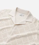 Camisa de manga corta color crema, confeccionada en tejido de punto con diseño de rombos calados y pequeños bordados florales en el centro de cada rombo. Posee cuello cubano y cierre frontal de botones.