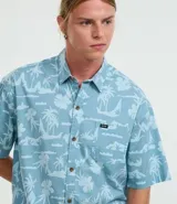 Camisa celeste con estampado de islas, palmeras y flores, de corte relajado, manga corta, cuello inglés, abotonada y con bolsillo en el pecho.