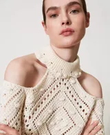 Sweater color crema de punto calado con motivo de rombos, cuello alto con volados y hombros descubiertos. Tiene mangas largas holgadas, ribetes acanalados, bajos festoneados y corte holgado.