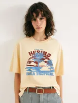 Camiseta de corte oversized color beige con estampa frontal estilo vintage que incluye palmeras y el texto 'Hering Baia Tropical'.