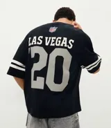 Remera deportiva de corte oversized, color negro, con estampado gráfico en la espalda del equipo Las Vegas Raiders de la NFL. Presenta cuello redondo, mangas cortas con detalles de rayas blancas y logo de la NFL en la parte superior trasera.