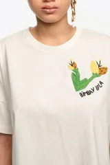 Remera de manga corta color crudo, con cuello redondo y diseño holgado. Presenta un bordado en el pecho con motivo verde y amarillo y la inscripción de la marca.