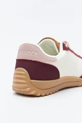 Zapatilla urbana de mujer con silueta retro, combinando paneles de cuero blanco y gamuza gris claro. Posee un talón trasero en gamuza negra y suela robusta de goma color caramelo con relieve.