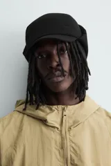 Gorra técnica de color negro con diseño de cinco paneles y orejeras integradas ajustables mediante una trabilla superior. Confeccionada en tejido acolchado, cuenta con ajuste elástico en la parte posterior.