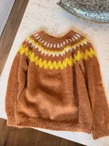 Sweater tejido a mano en mohair, de cuello redondo y manga larga. Presenta un diseño de canesú con patrón geométrico en tonos amarillo y blanco sobre una base de color marrón.