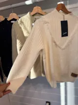 Sweater de punto color blanco, con cuello alto y detalle de escote en V con terminaciones desflecadas. Presenta mangas largas con puños acanalados y un diseño de calce holgado.