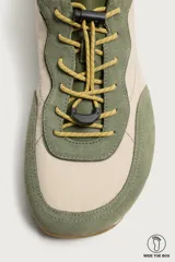 Zapatilla deportiva barefoot fabricada en combinación de piel y tejido, en tonos verde oliva y beige, con cordones elásticos amarillos y suela de goma flexible.