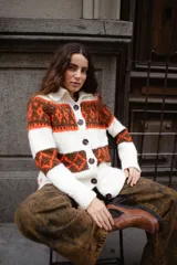 Saco tejido a mano con diseño jacquard en tonos rojo, naranja y marrón. Presenta cuello tipo polo, cierre frontal con botones grandes, mangas largas y tajos laterales en el ruedo.