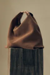 Cartera tipo hobo de cuero color camel, con diseño holgado y cierre superior. Presenta un detalle metálico en la parte superior y el logo de la marca grabado en el frente.