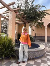 Camisa de manga larga color naranja con cuello grande en contraste color rosa y detalle de volados en el borde. Presenta un diseño con botones frontales y un detalle de tela rosa en la parte inferior.