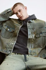 Campera de denim de algodón rígido, color verde azulado desgastado, con cuello, cierre frontal de botones metálicos y dos bolsillos superiores con solapa y botón. Presenta puños largos abotonados y trabillas ajustables con botones en los laterales.