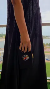 Cartera tipo clutch de gamuza negra, de forma rectangular, con cierre metálico dorado en el lateral y detalle de aplique floral brillante con piedras de colores en el centro.