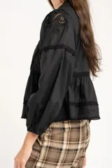 Blusa negra de algodón con diseño de broderie y calados en el pecho y hombros. Presenta corte oversize, mangas largas con puños y un detalle de volado en la cintura.