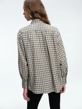 Camisa de corte clásico con estampado de cuadros vichy en tonos azul y blanco. Presenta cuello camisero, cierre frontal con botones ocultos y mangas largas con puños abotonados. Incluye un detalle de broche metálico dorado en el cuello.