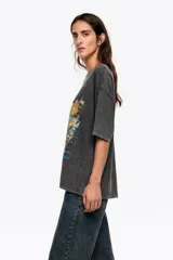 Remera de algodón, color gris oscuro con efecto gastado (acid wash), corte oversize y escote redondo. Presenta un estampado frontal artístico con una ciudad, flores amarillas y una bota de lluvia verde.