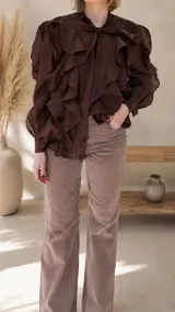 Camisa de color marrón chocolate, confeccionada en tela liviana y fluida. Presenta un diseño con múltiples vuelos frontales, cuello con lazo para anudar y mangas largas abullonadas con puños ajustados.