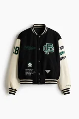 Campera estilo varsity con cuerpo negro y mangas en contraste color crema. Presenta múltiples parches y bordados decorativos en el frente y las mangas, cuello, puños y ruedo acanalados a rayas, y cierre frontal con botones a presión.