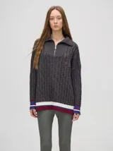 Pullover de algodón color beige, con tejido en ochos, cuello polo con cierre frontal y puños a rayas de colores (blanco, bordó y amarillo). El diseño es holgado y cae por debajo de la cadera.