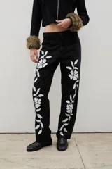 Pantalón de denim negro de corte recto con estampado floral blanco en ambas piernas. Cuenta con cierre frontal y bolsillos laterales.