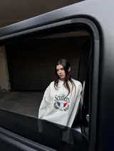 Buzo de corte boxy color crudo, con cuello redondo y mangas largas de hombros caídos. Presenta un estampado frontal con el logo de la marca, un escudo con los colores de la bandera italiana y detalles de laureles.