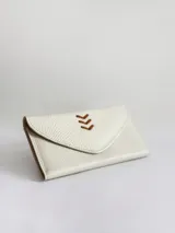 Billetera de cuero vacuno color crudo con textura granulada. Presenta un diseño tipo sobre con cierre de broche y un detalle decorativo de espiga bordado en cuero color suela en la solapa frontal.