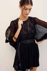 Vestido corto negro con transparencias, mangas largas amplias y detalles de bordados florales con lentejuelas en el frente. Cuenta con un lazo ajustable en la cintura y falda fluida.