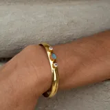 Brazalete rígido dorado con tres piedras incrustadas: una labradorita ovalada en el centro y dos piedras de la luna redondas a los lados.