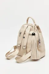 Mochila de símil cuero texturizado color beige, con asa superior, tiras regulables para la espalda y cierre principal metálico. Presenta un detalle trenzado vertical en el frente y un accesorio con flecos y cuenta negra en el lateral.