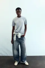 Camiseta gris de cuello redondo y manga corta, con un estampado en relieve que dice "NEW YORK CITY" y "BROOKLYN". El tejido tiene un sutil brillo.