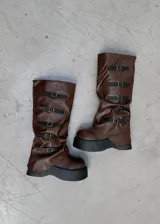 Botas bucaneras de cuero vegano color marrón, con caña arrugada y plataforma gruesa negra. Presentan cinco tiras ajustables con hebillas doradas a lo largo de la caña.