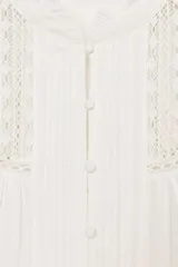 Camisa blanca de cuello redondo con volantes, manga larga con puños elásticos y cierre frontal con botones forrados. Presenta detalles de puntilla y jaretas en el delantero.