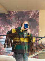 Campera de lana italiana con diseño de rayas horizontales en tonos verde, amarillo y marrón. Presenta flecos largos de gamuza en las mangas, cierre frontal y bolsillos laterales. La prenda se encuentra completamente forrada.