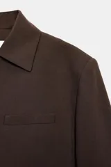 Blazer de corte estructurado en color marrón chocolate, con cuello solapa, hombros marcados y cierre frontal cruzado con botón oculto. Presenta un bolsillo superior tipo ojal y un diseño de bajo redondeado.