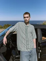 Polo de punto verde con rayas verticales blancas, corte relajado, cuello tipo camisa, cierre con botones y mangas cortas.