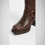 Bota de caña alta confeccionada en cuero vacuno, con diseño de punta cuadrada y plataforma. Presenta un taco cuadrado de madera, cierre lateral interno y una tira decorativa con herraje metálico circular en el tobillo.