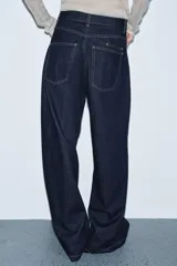 Jeans de tiro alto con pierna ancha, confeccionados en denim azul oscuro. Presentan cinco bolsillos, trabillas en la cintura y un cinturón ajustable del mismo tejido con hebilla metálica. Incluyen pespuntes marcados a tono y cierre frontal con cremallera y botón.