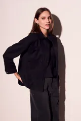 Sobrecamisa negra de estilo shacket, confeccionada en tejido de gamuza sintética. Presenta cuello camisero, cierre frontal de botones y dos bolsillos de parche con solapa en el pecho. El calce es oversize y las mangas son largas.