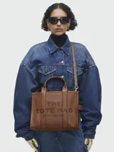 Bolso tipo tote de cuero negro, modelo The Leather Medium Tote Bag de Marc Jacobs. Presenta doble asa superior, correa ajustable para el hombro y el texto grabado "THE TOTE BAG MARC JACOBS" en el frente.