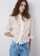 Blusa de tejido semitransparente color crudo, con diseño recto, cuello alto con volados, mangas largas abullonadas con puños abotonados y detalle de bordado floral en toda la prenda. Cuenta con cierre frontal de botones ocultos y ribete con volado en la botonera.