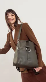 Mochila de tamaño mediano, color beige claro, con cuerpo principal de tela impermeable y detalles en simil cuero liso en la parte inferior y correas. Presenta un asa superior corta y correas ajustables para llevar al hombro o cruzada. Cuenta con un bolsillo frontal con cierre y un detalle decorativo de correa con hebilla metálica rectangular.