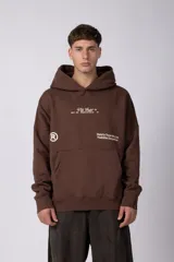Buzo canguro color marrón con capucha, confeccionado en frisa de algodón. Presenta un diseño gráfico estampado en el frente con texto y logo circular, bolsillo tipo canguro y puños acanalados.