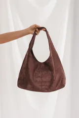 Bolso tipo hobo de gamuza color marrón, con diseño amplio y asa integrada al cuerpo del bolso.