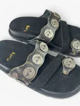 Sandalias planas de cuero negro con diseño de tiras anchas decoradas con apliques metálicos circulares grabados. Presentan una tira separadora para el dedo y plantilla forrada en cuero.